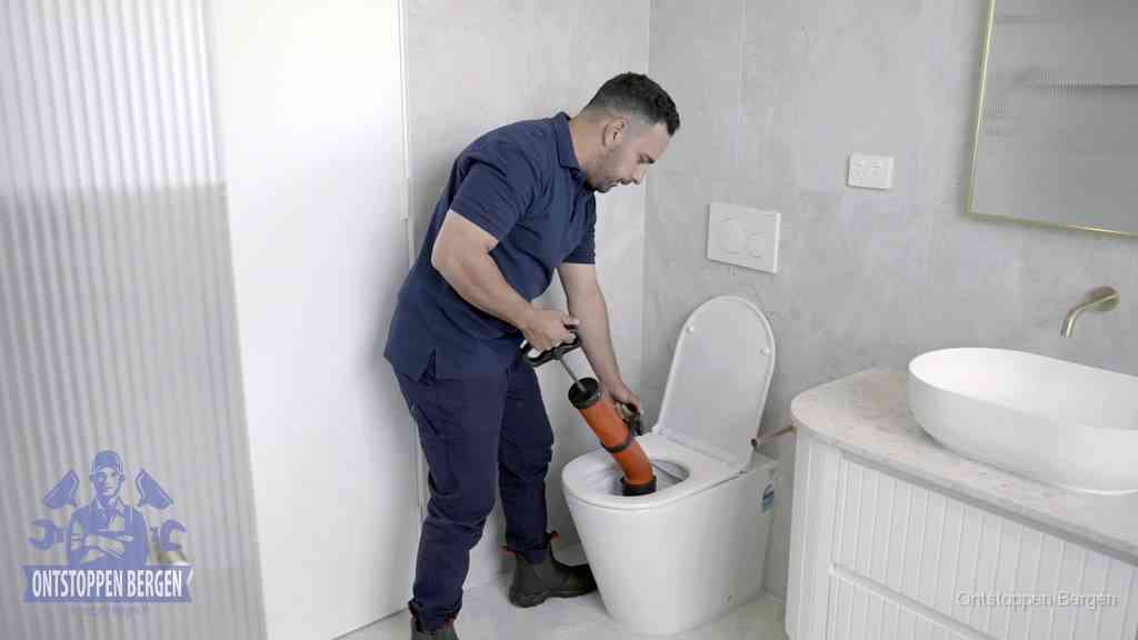 Een monteur buigt over een wandcloset en duwt met de hand een persluchtontstopper in de toiletpot om de verstopping handmatig los te maken in Bergen