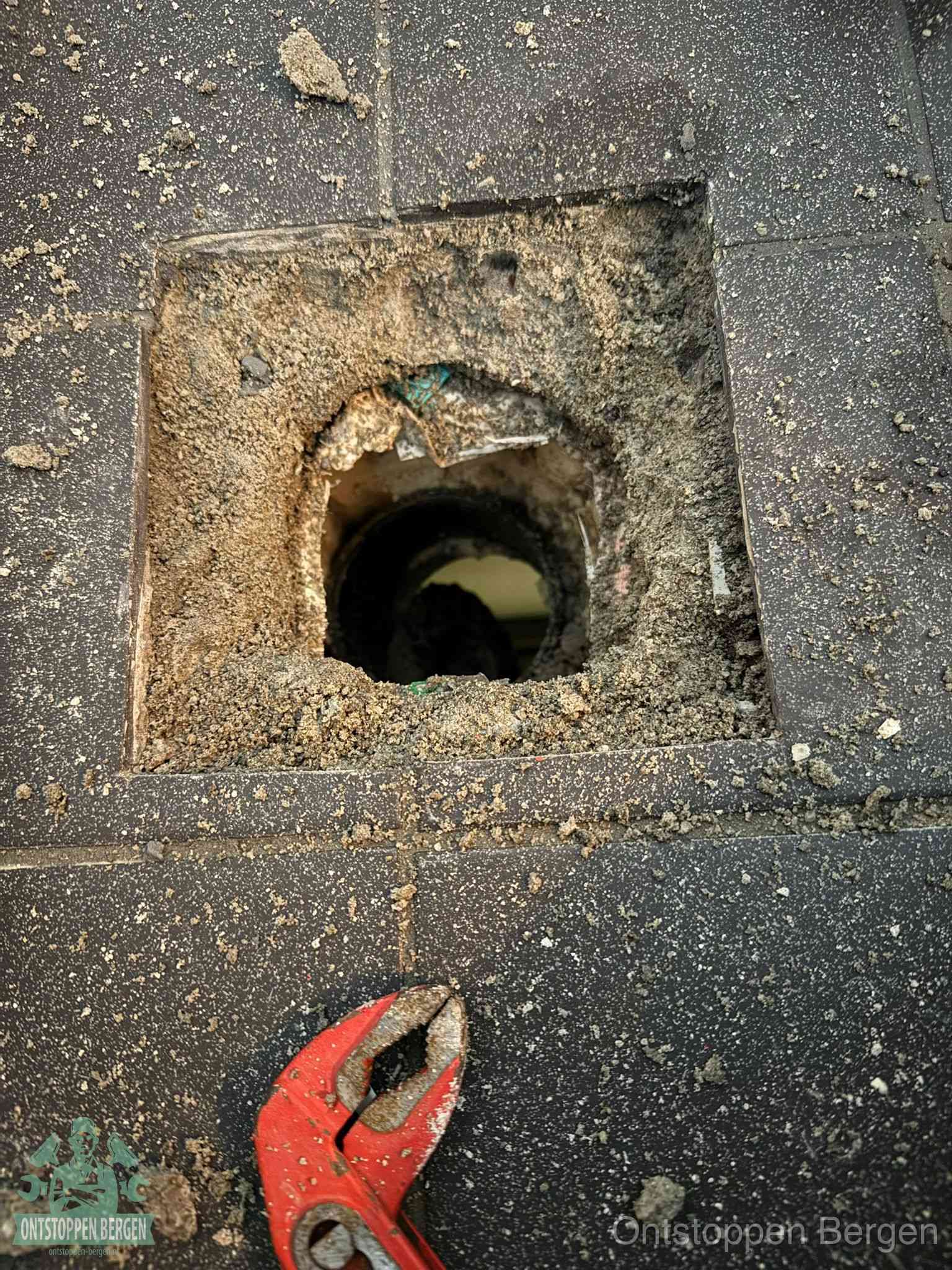 Een geopend rioolputje in een betontegelvloer met losgemaakt rooster en een zichtbare ronde afvoerbuis, het achterhalen waar de afvoerverstopping zit in Bergen