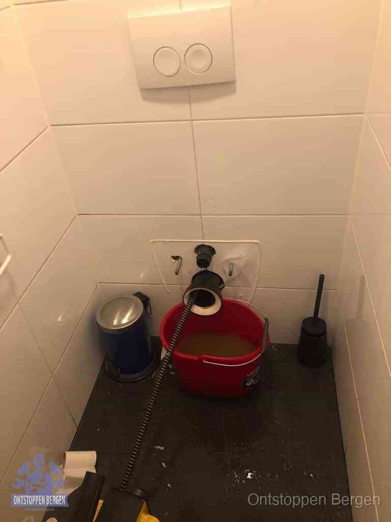 Een ontstopte wand-wc met verwijderde toiletpot waar een ontstoppingsveer in de wanduitgang verdwijnt, het achterhalen waar de verstopping zit in Bergen