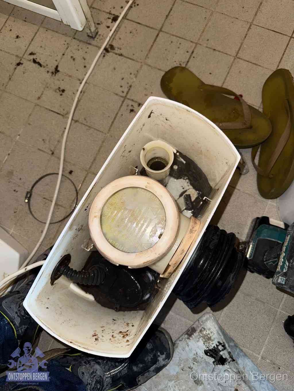 Een gedemonteerde toiletpot op de vloer met ontstoppingsveer en gereedschap ernaast, het verwijderen van een vastzittend voorwerp uit de wc in Bergen