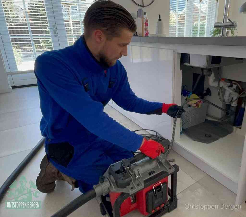 Een monteur met rode handschoenen knielt in een keuken en voert een elektrische ontstoppingsveer in tijdens het professioneel afvoer ontstoppen in Bergen